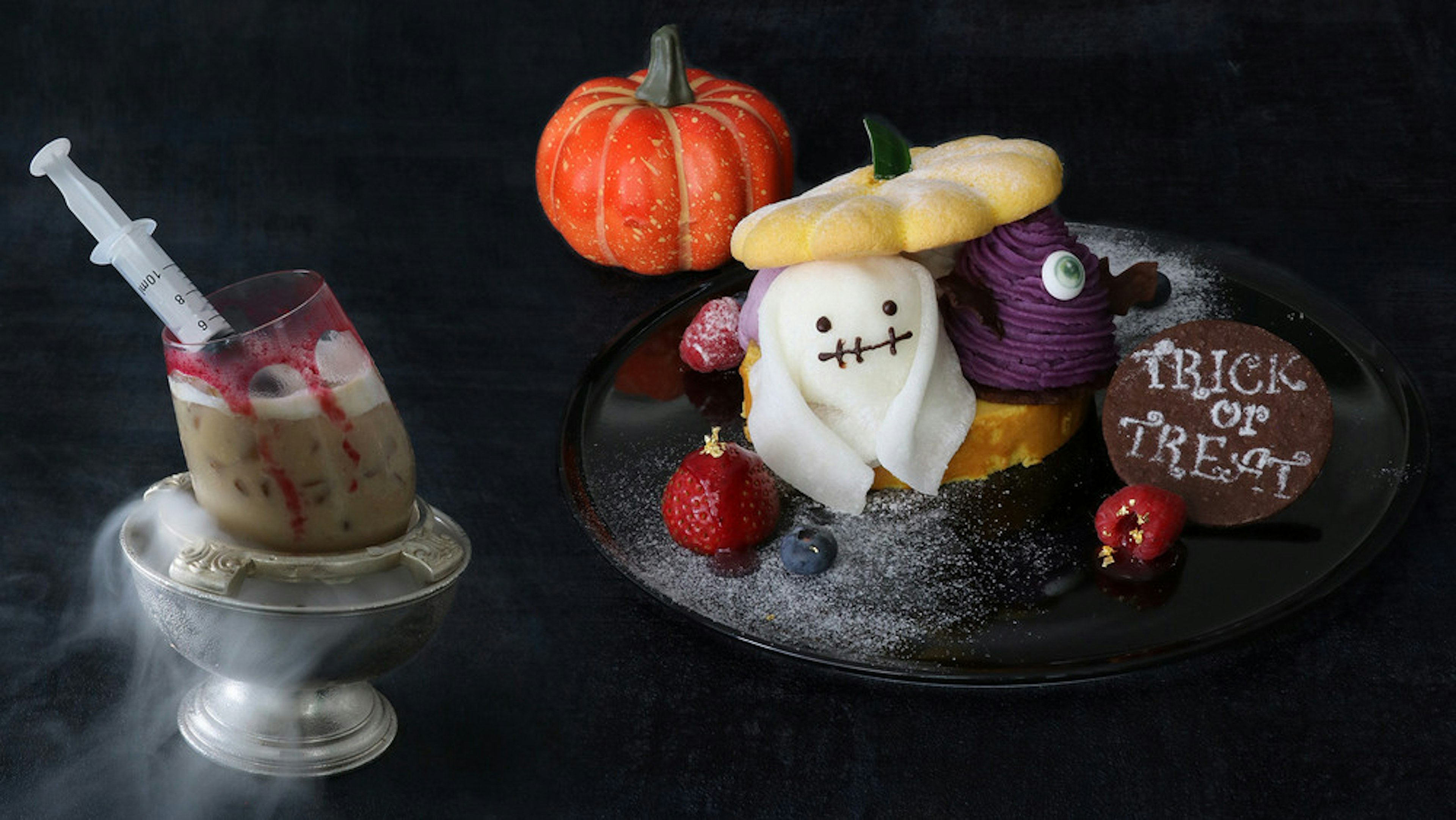 黒い背景にハロウィンをテーマにしたデザートが美しく盛り付けられたプレートがある。ゴーストの形をしたスイーツや、紫色のモンブラン風クリーム、かぼちゃのような形のパイが飾られて、隣には「Trick or Treat」と書かれたクッキーも添えられている。左側には注射器が刺さった不気味なドリンクが銀のカップに入っており、ハロウィンらしいユニークでちょっと怖い雰囲気が漂っている画像。