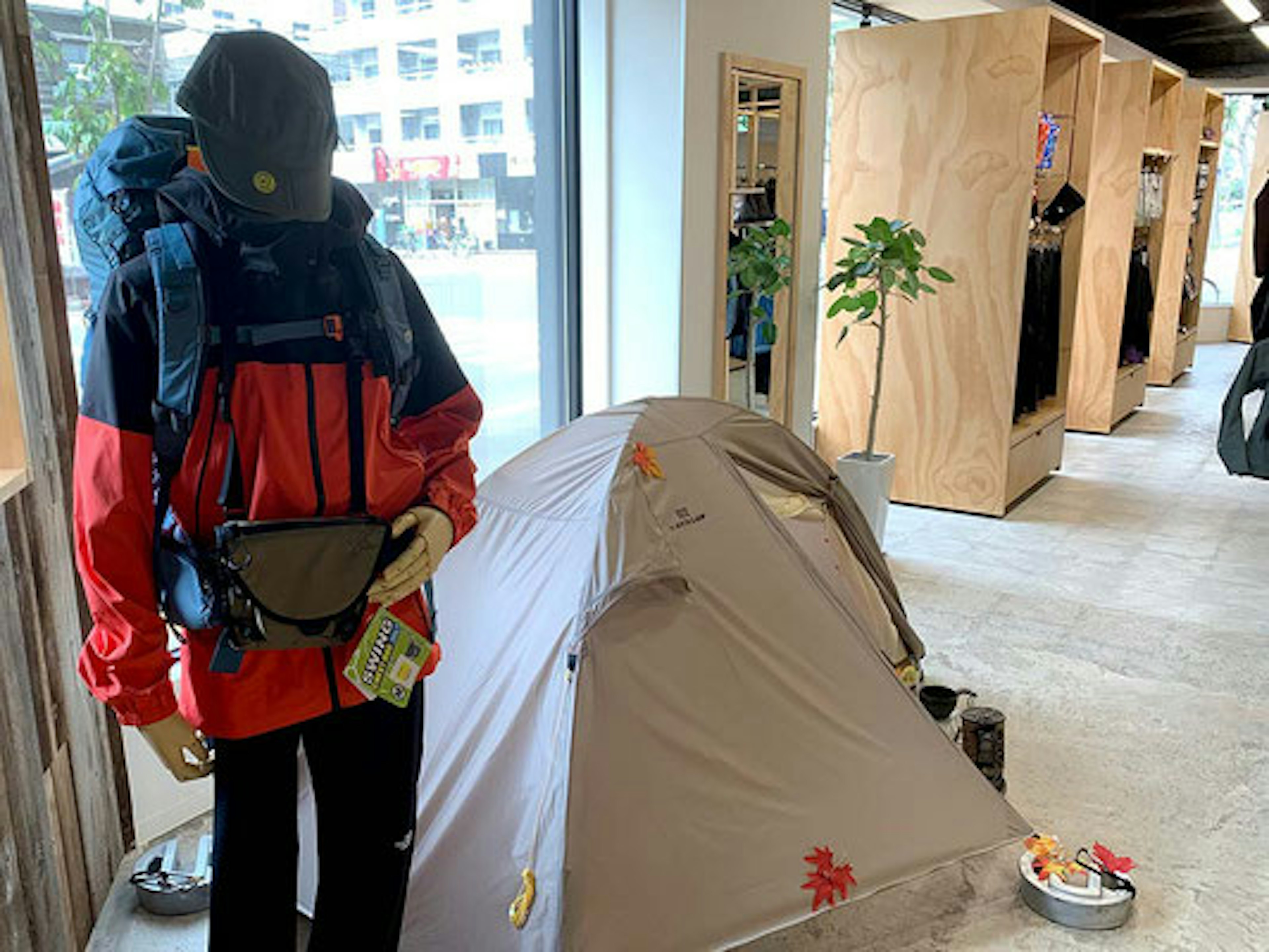 アウトドア用品店の店内ディスプレイで、マネキンが赤と黒のジャケットを着てリュックサックを背負い、帽子をかぶっている。マネキンの隣にはキャンプ用のテントが設置され、地面には小さなクッキングセットや他のキャンプ用品が配置されている。