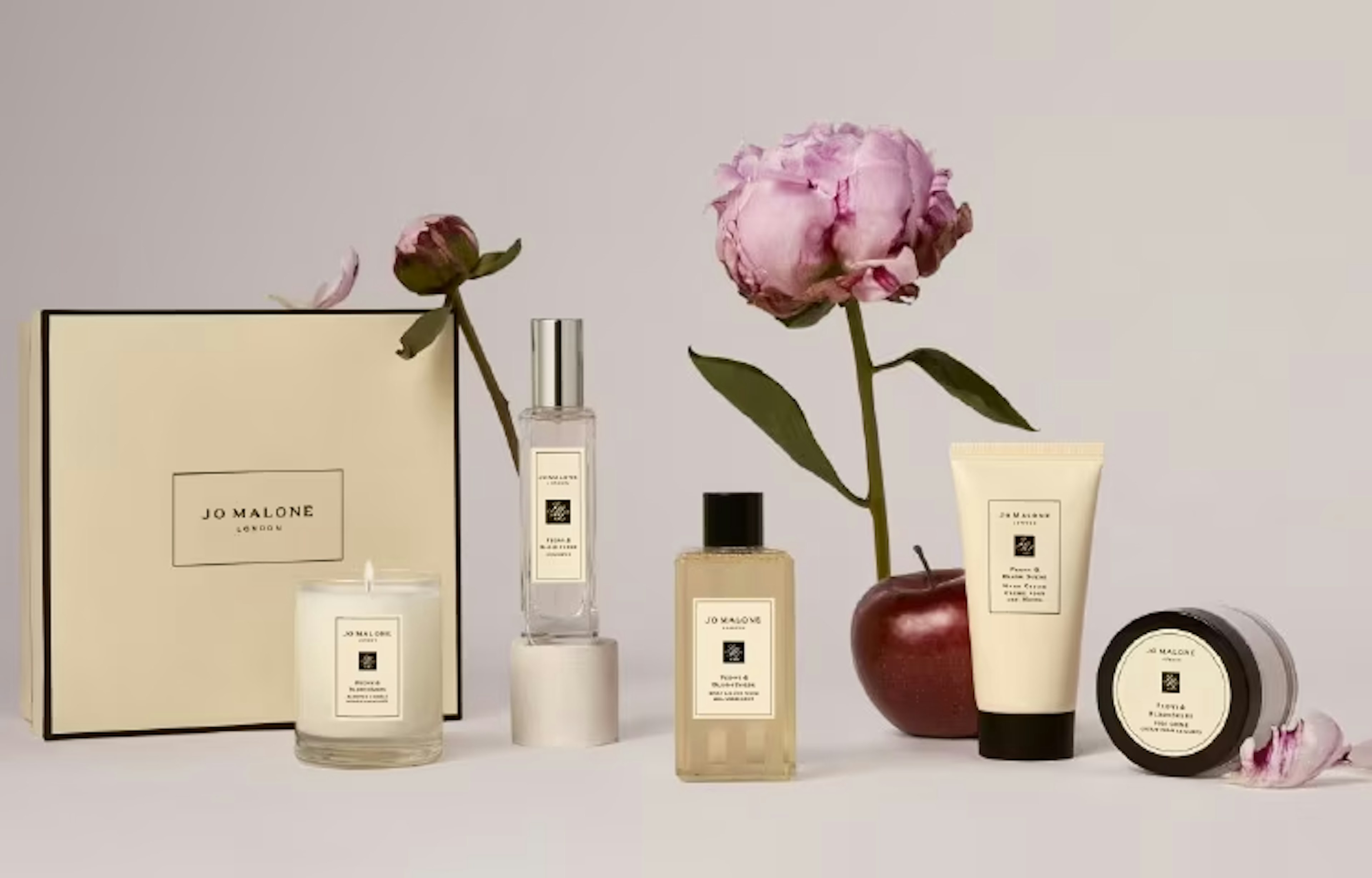 白い背景の上に、JO MALONEの製品が並べられた美しいディスプレイ。左から、JO MALONEのロゴが入ったボックス、キャンドル、ボトルのフレグランス、透明ボトルのリキッドソープ、ハンドクリーム、小さなジャーのバームが配置されている。ピンクの芍薬の花と赤いリンゴがアクセントとして置かれ、シンプルでエレガントな雰囲気を演出している画像。