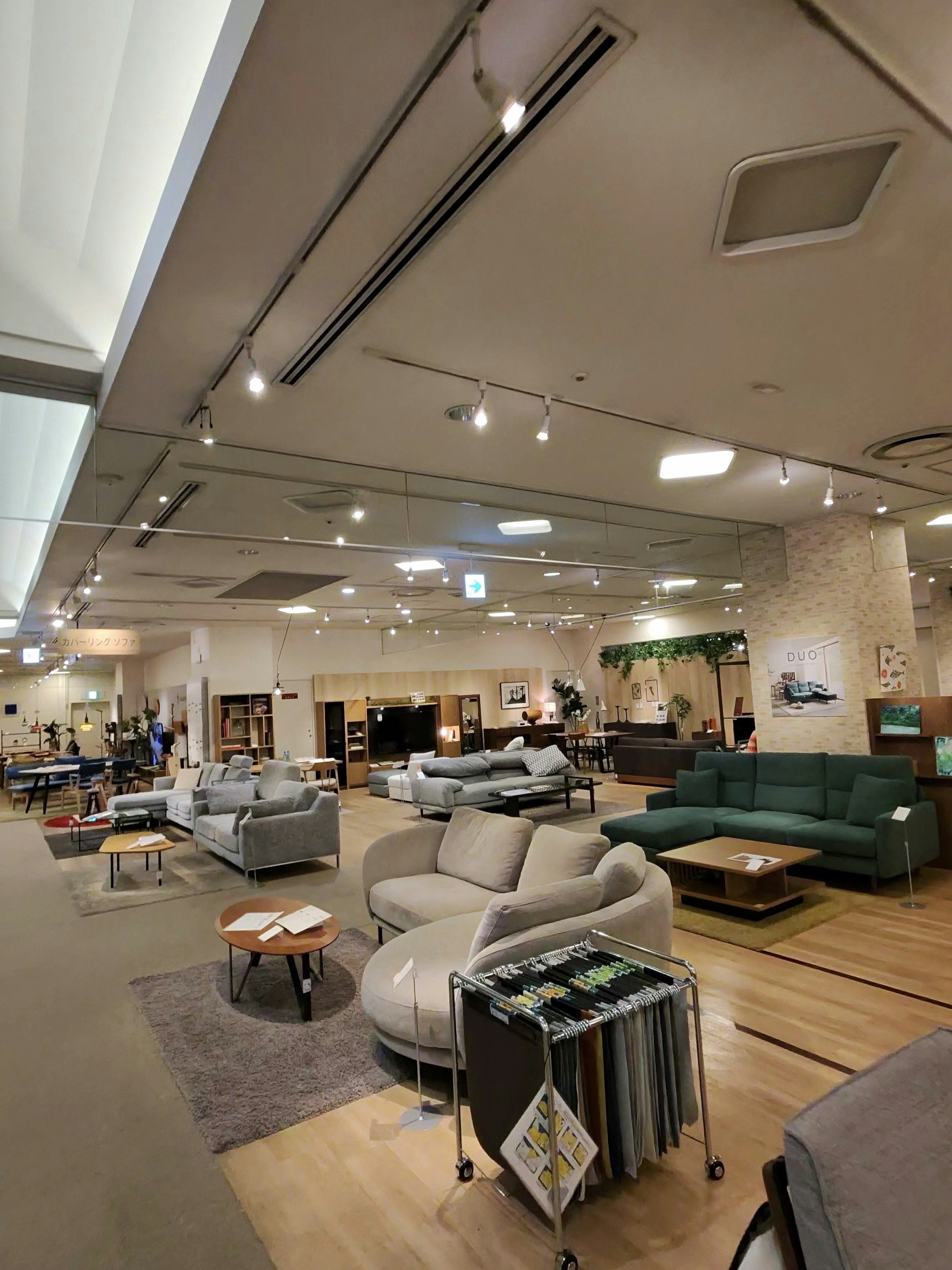家具展示スペース内のさまざまなスタイルのソファとテーブルが配された広い店内。左側には灰色のファブリックソファ、クッションがいくつかあり、右側には緑色のソファセットが目立つ。部屋の中央にはカラフルなカーペットが敷かれ、さまざまなインテリアアクセサリーが展示されている。