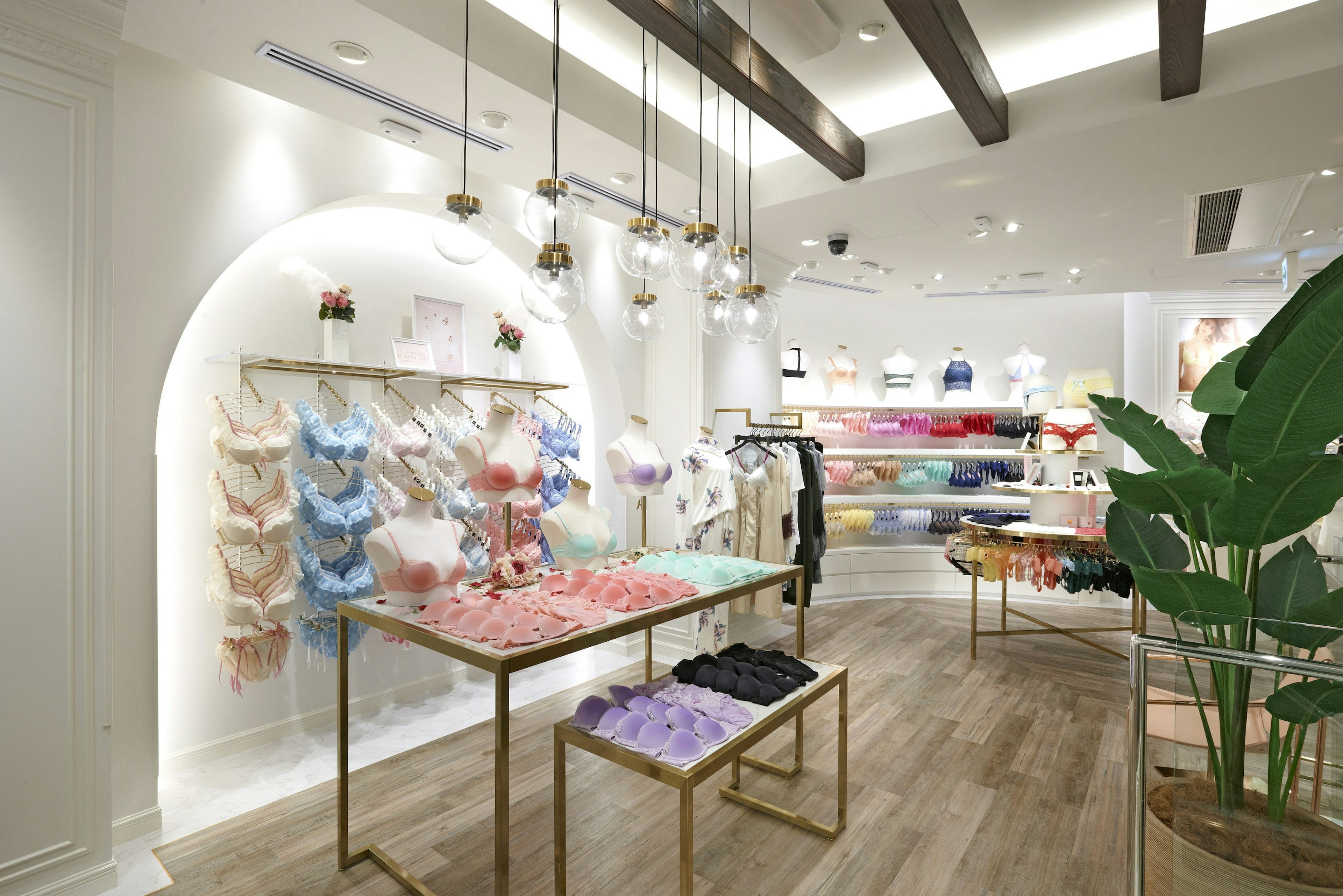 下着専門店。店内には様々な色とデザインのブラジャーが美しく展示されており、中央のテーブルには色別に整理された下着が並べられている。天井から吊り下げられた現代的な照明と、店内の大型植物が洗練された雰囲気を演出している。