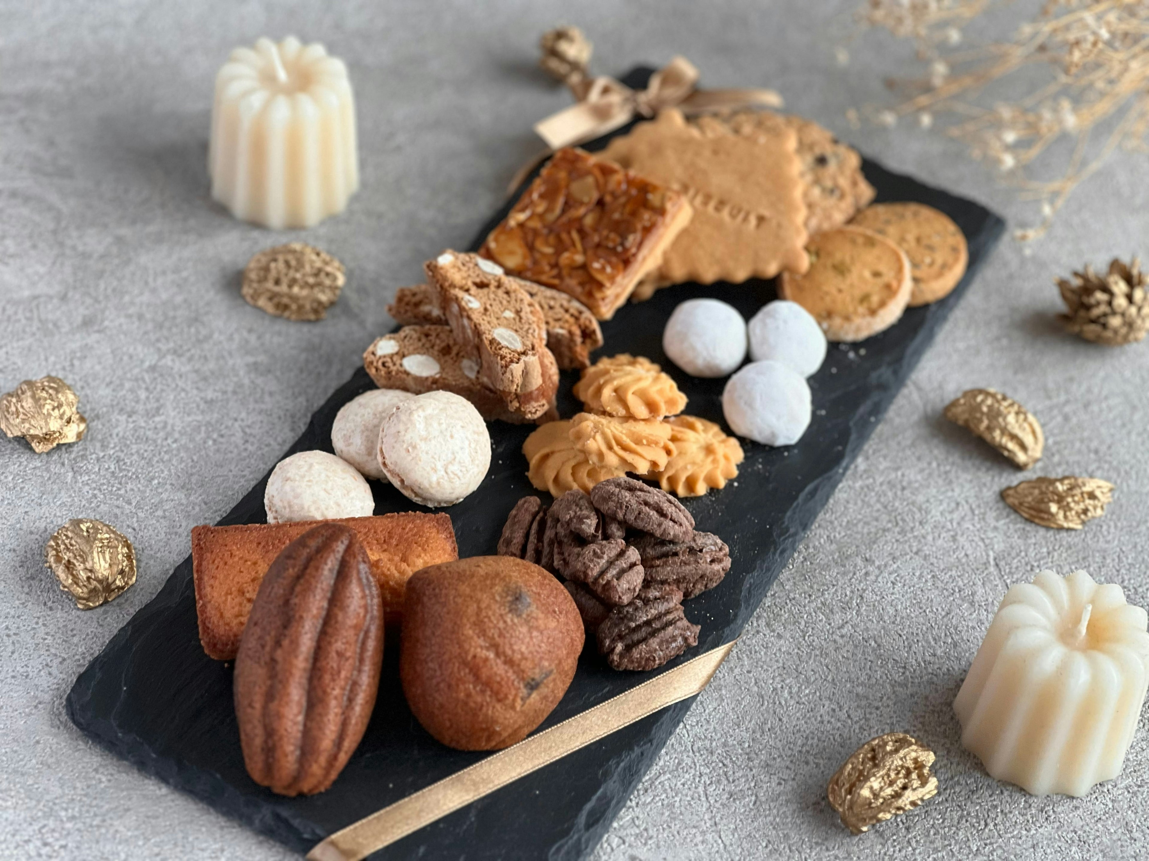 黒いプレートに盛り付けられた様々な焼き菓子が並び、周囲にキャンドルや松ぼっくりが装飾されている。