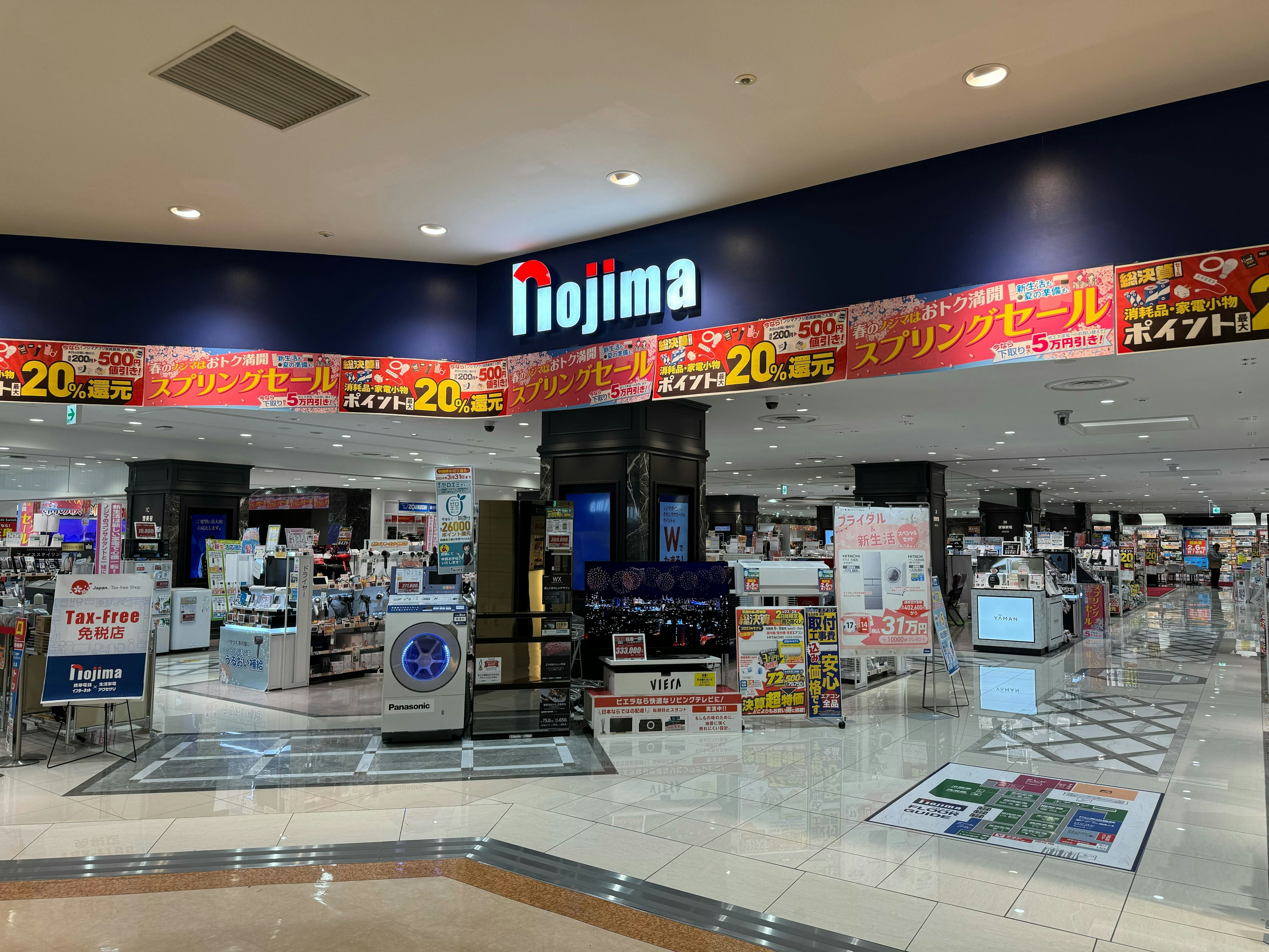 ノジマ電器店の広々とした店内入口。多様な電化製品がディスプレイされており、大きな赤い看板でさまざまなセール情報がアピールされている。店内は明るく清潔感があり、入口付近には税込み価格での販売を示す「Tax-Free」のサインが設置されている。