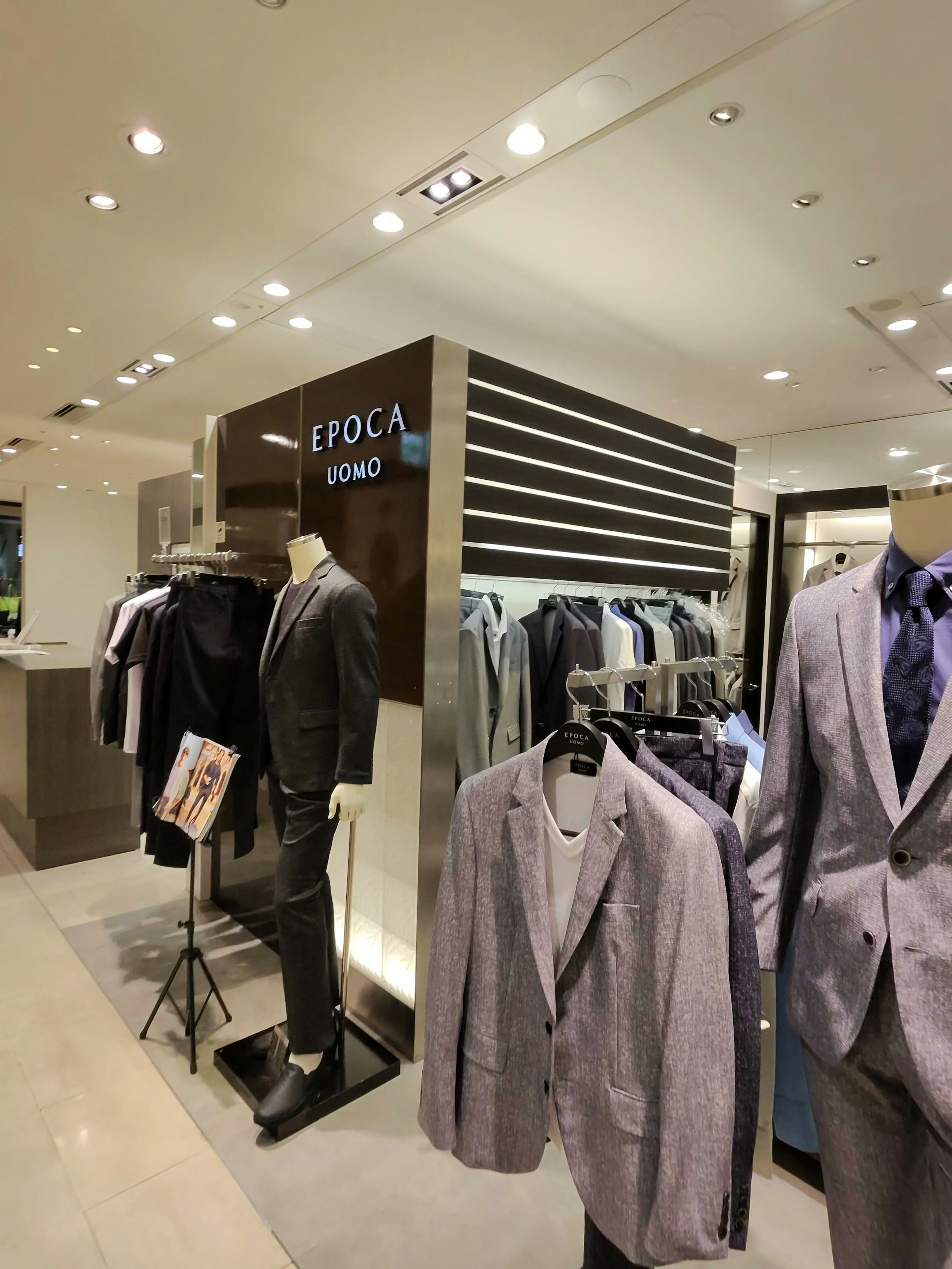 上質なスーツとジャケットが展示されており、一体型のマネキンがスタイリッシュなアンサンブルを着用している。背景には「EPOCA UOMO」と明記されたブランドのサインがあり、モダンで洗練された店の雰囲気。