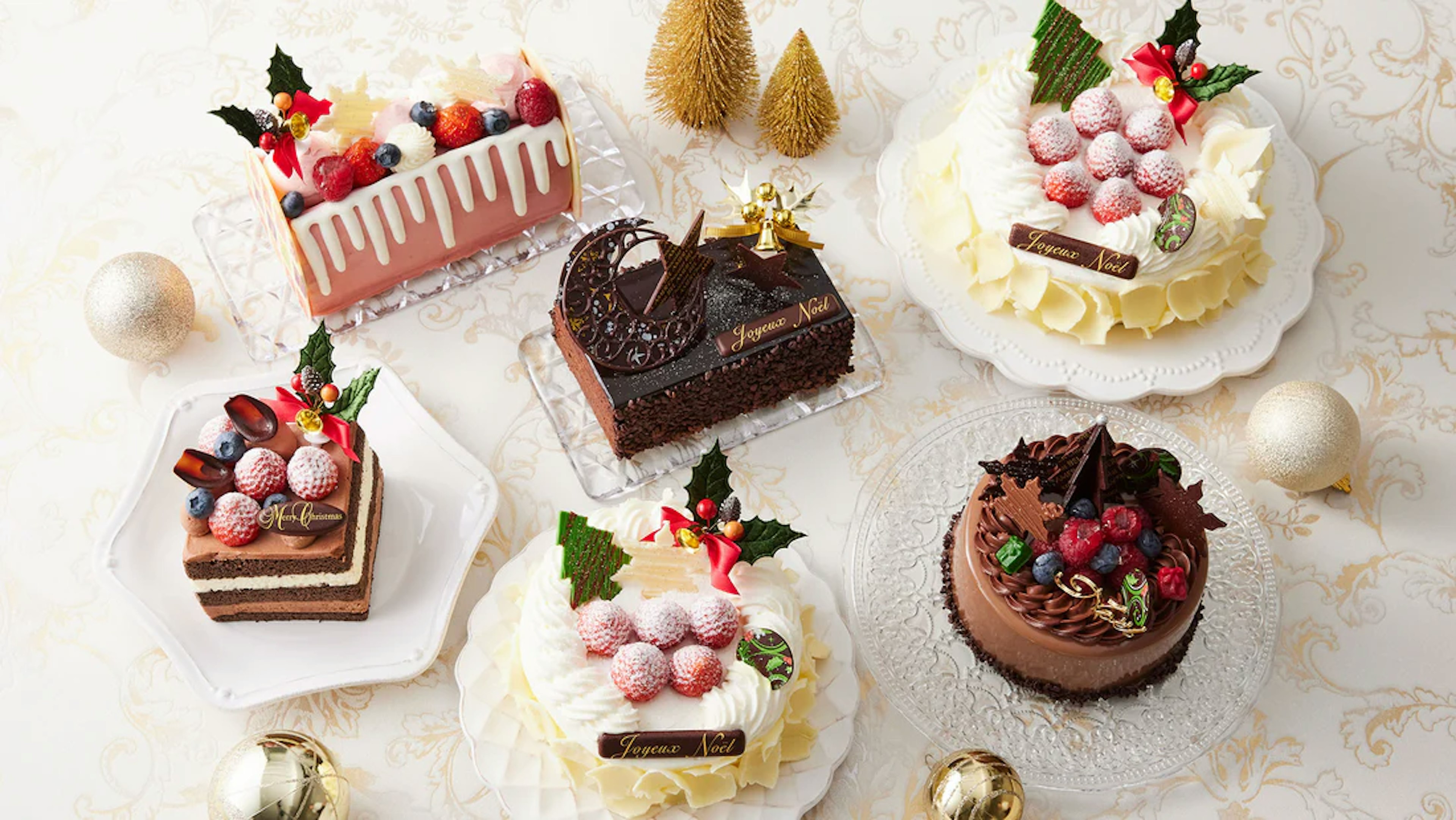 色とりどりのクリスマスケーキが並んでいる。いちごやベリーでデコレーションされたホワイトクリームのケーキや、チョコレートでコーティングされたリッチなケーキ、そしてホワイトチョコレートのドリップが美しい長方形のケーキなど、さまざまなデザインのケーキが並べられている。どのケーキにも「Joyeux Noël」と書かれたチョコレートプレートが飾られ、クリスマスの華やかな雰囲気が表現されている。背景にはゴールドのボールオーナメントやクリスマスツリーの小さな装飾があり、全体的にエレガントなクリスマスの演出がされている画像。