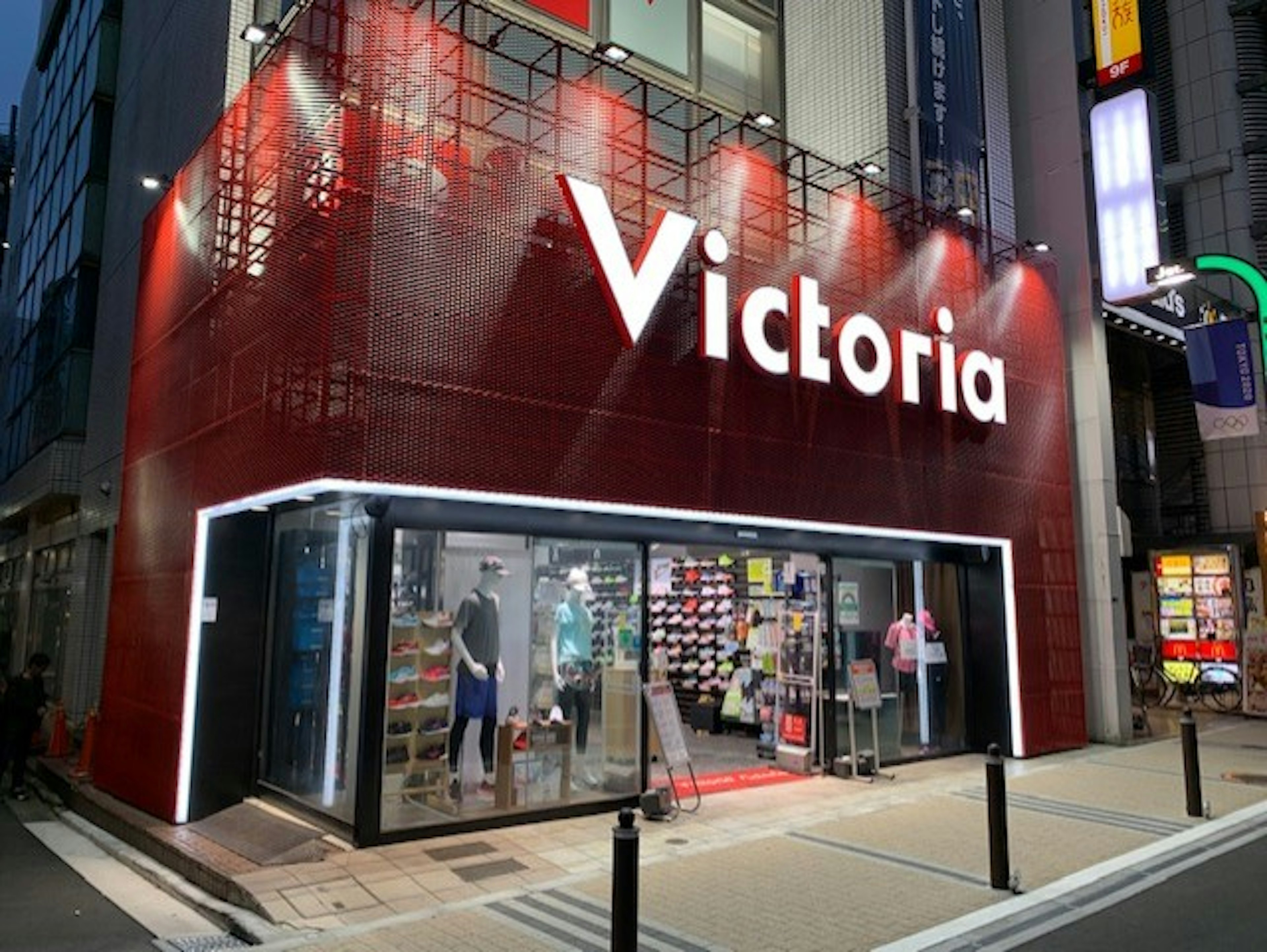 「Victoria」という看板を持つスポーツ用品店の入り口。店の外観は赤い格子状のデザインで、店名の「Victoria」が白く明るく照らされている。ガラス張りの入り口からは、スポーツウェアやシューズがディスプレイされている。店内は明るく、商品がきれいに並べられている。背景には他の建物や看板も見え、都会の雰囲気が感じられる。