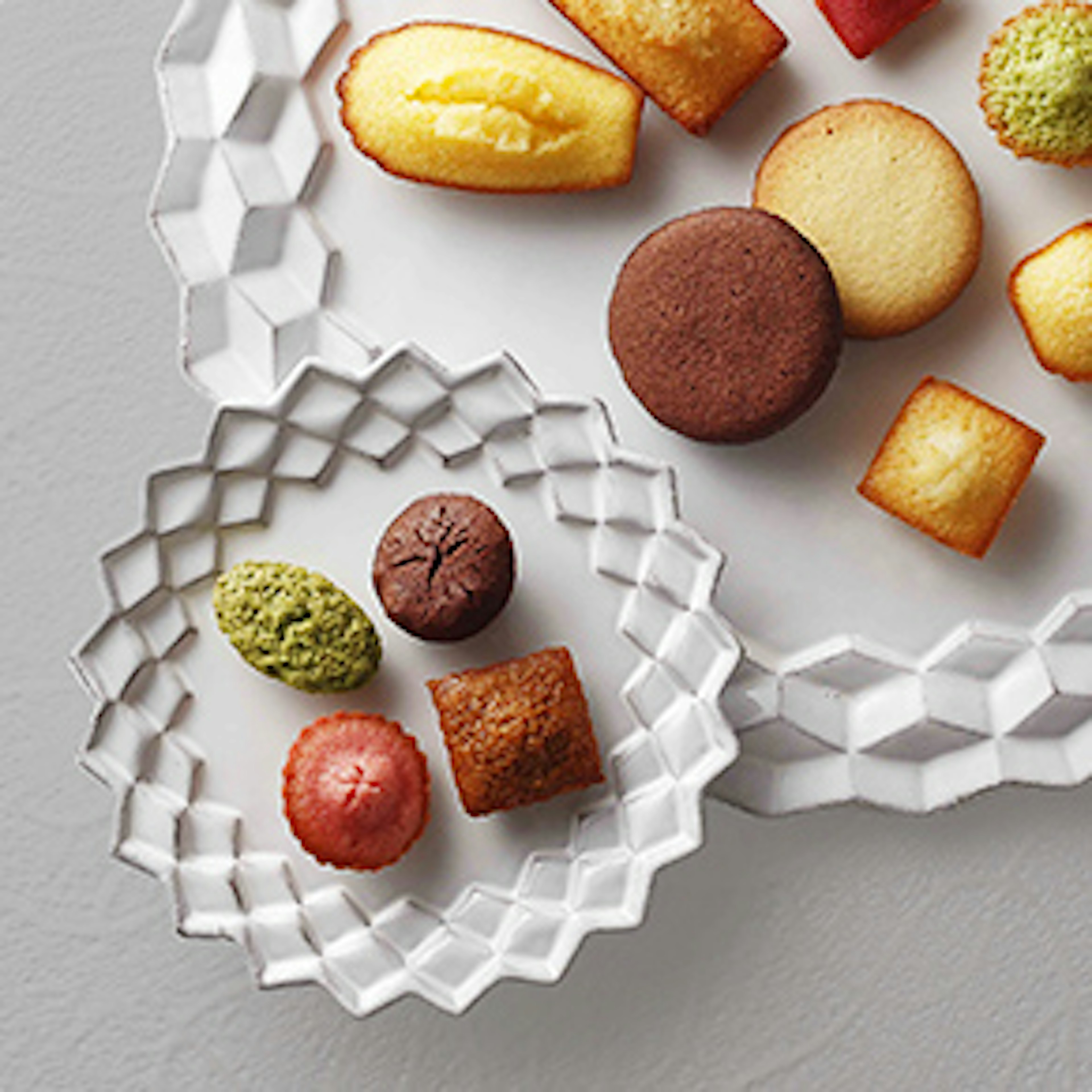 二つの幾何学模様のプレートに、小さな焼き菓子が色とりどりに並べられている。大きなプレートにはフィナンシェやマドレーヌが配置されており、小さなプレートには抹茶やココアなどの焼き菓子が載せられていて、シンプルで洗練されたデザインが印象的な画像。