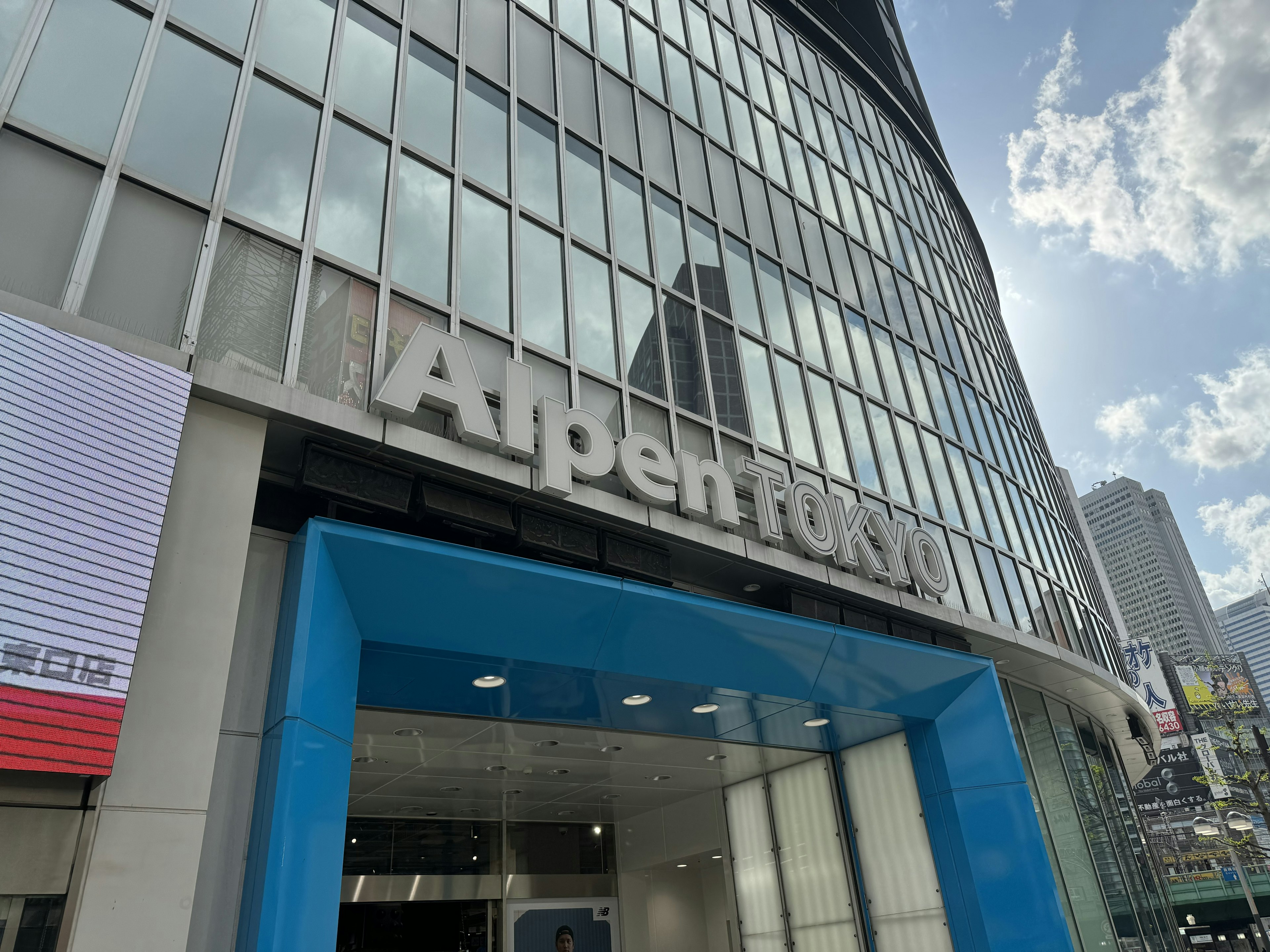 大きなガラス窓とモダンな建築スタイルが特徴的なビルで、店名「Alpen TOKYO」がファサードに大きく掲示されている。エントランスは鮮やかな青のアクセントカラーで装飾されている。