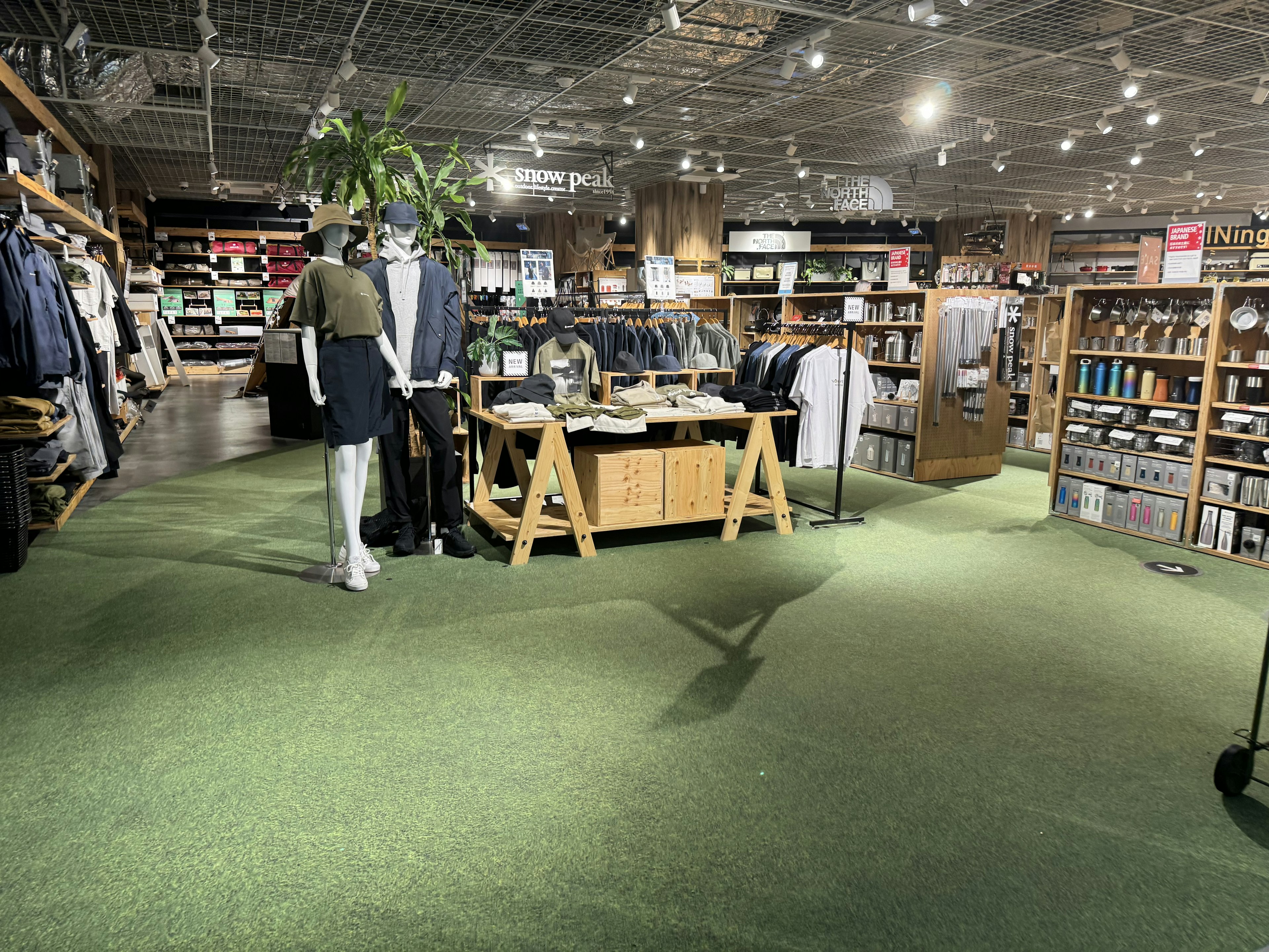 Alpen Tokyoのアウトドアコーナー。床には緑のカーペットが敷かれ、服、靴、キャンプ用品などが豊富に陳列されている。背景には「Snow Peak」と「The North Face」のロゴが見える