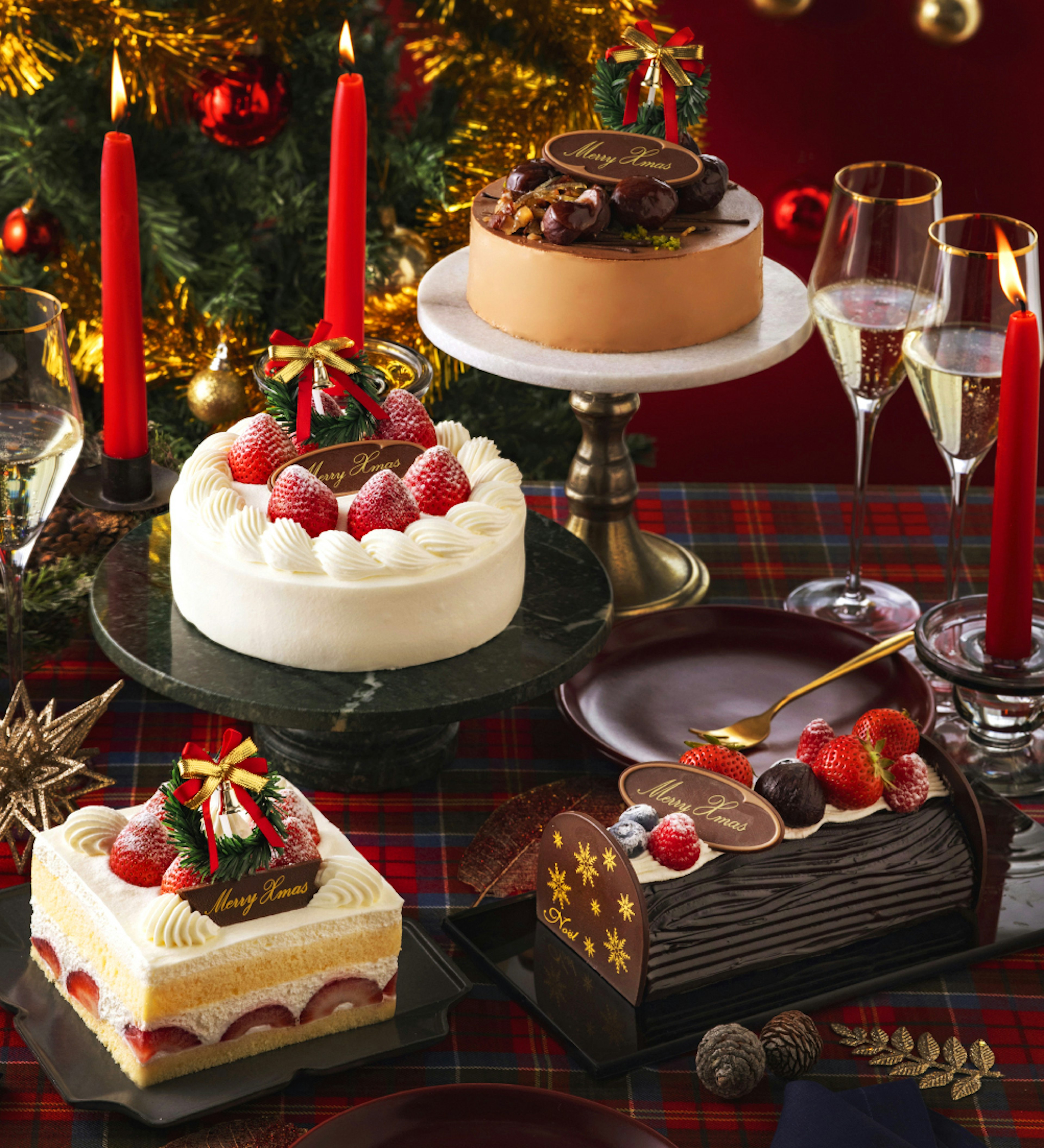 クリスマスをテーマにしたテーブルセッティング。中央には、4つのクリスマスケーキが飾られており、赤いキャンドルとクリスマスツリーが背景に見える。丸いケーキは白いクリームでデコレーションされ、いちごがたっぷりとトッピングされている。チョコレートケーキにはナッツが飾られており、他のケーキもいちごやクリームで彩られている。テーブルは赤と緑のチェック柄のクロスが敷かれており、クリスマスらしい雰囲気が漂っている画像。