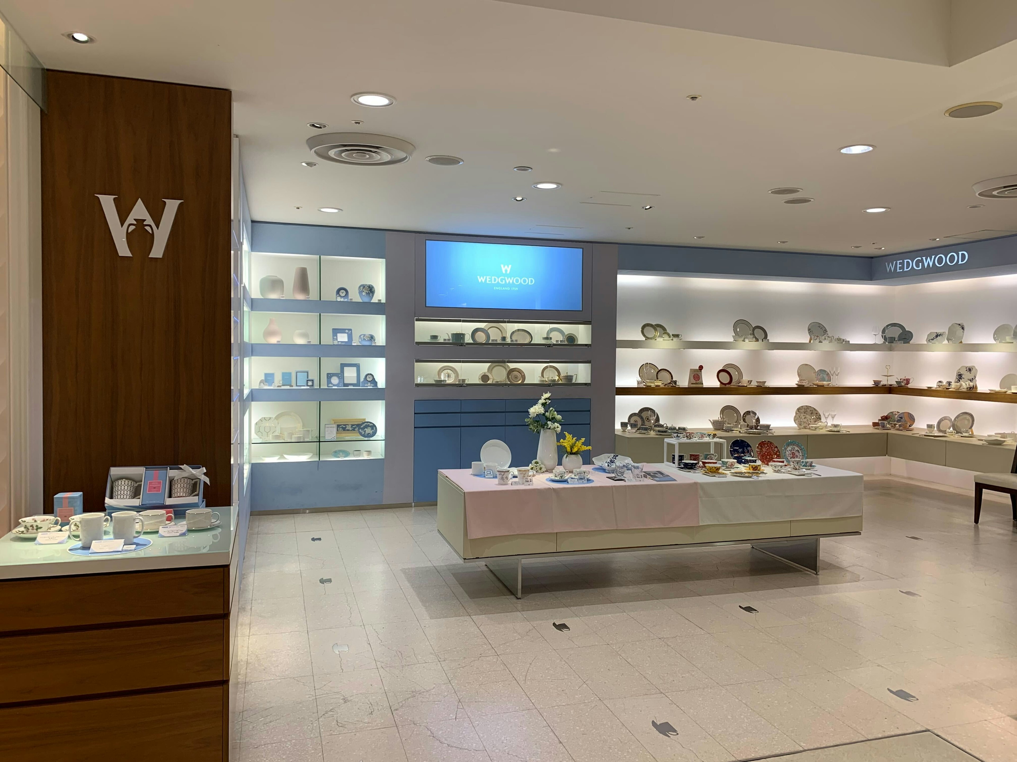 WEDGWOODの店内。壁に沿って様々なデザインのプレートやカップが美しくディスプレイされています。中央のテーブルには、花瓶に入った黄色と白の花が飾られており、その周りに多くの食器が並べられています。