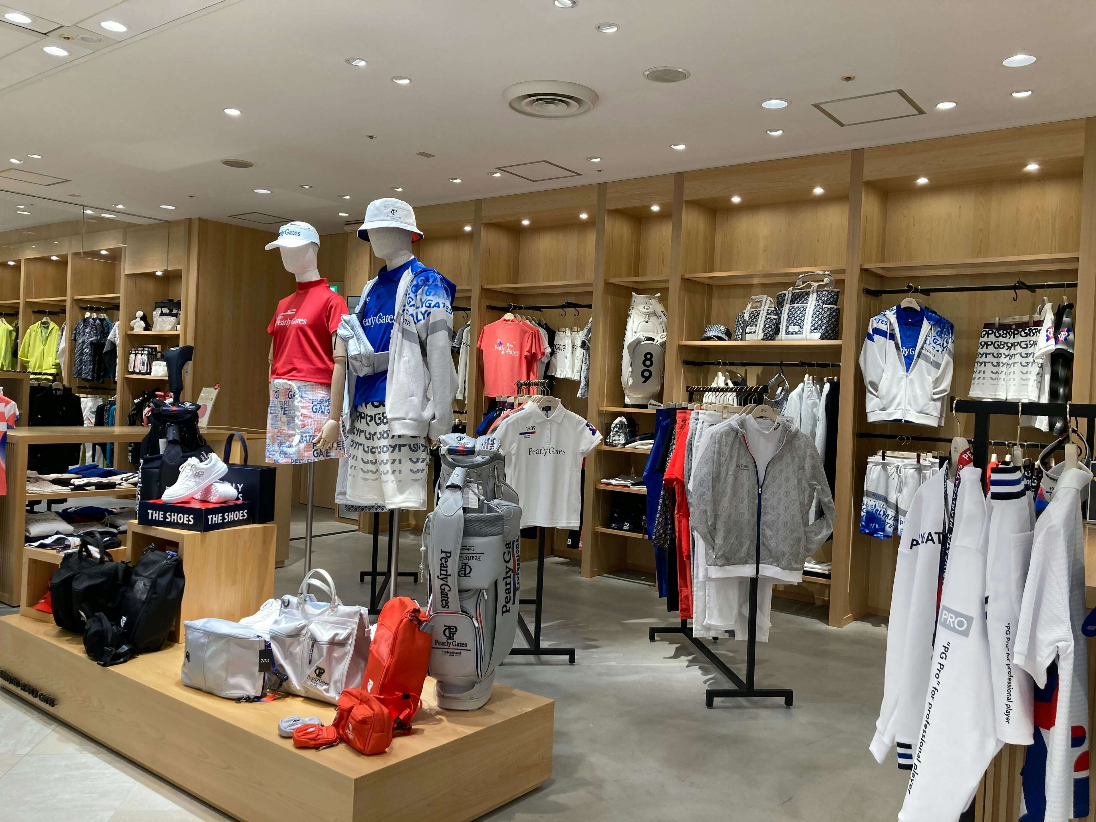 ゴルフウェア専門店の店内で、色とりどりのスポーツウェアが展示されている。マネキン二体がカジュアルなゴルフ服を着用しており、周囲にはゴルフバッグ、シューズ、その他のアクセサリーが配置されている。店内は木目調の内装で明るく、洗練された雰囲気が漂っている。