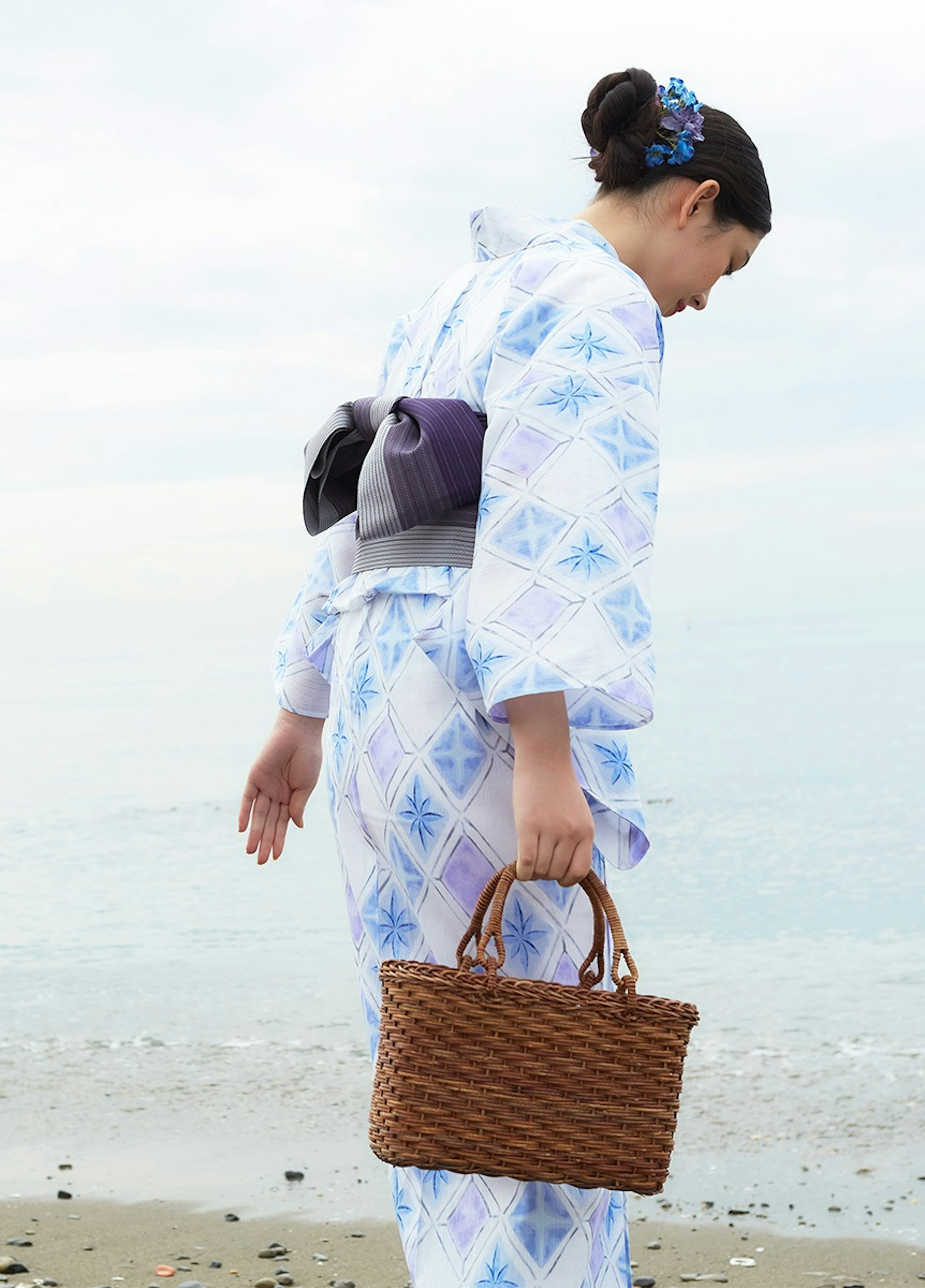 海辺で青い柄の浴衣を着た女性が、籐のかごバッグを持っている。