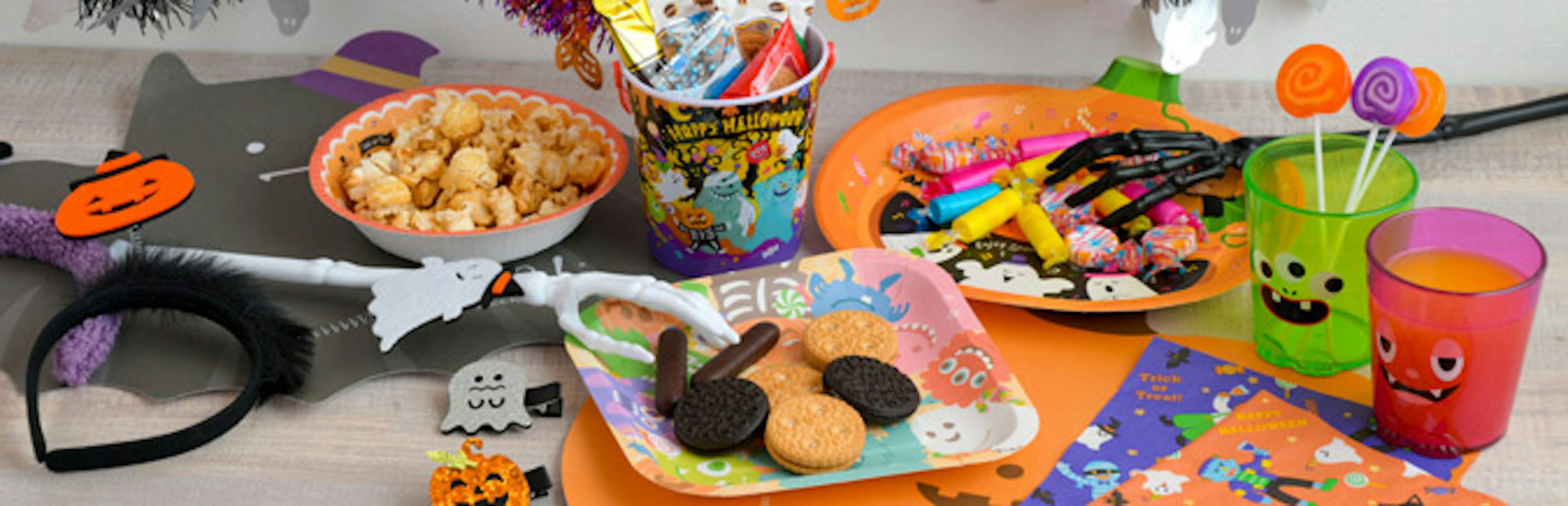ハロウィンのテーマに沿ったお菓子や装飾品がテーブルに並べられている画像。ポップコーンやクッキー、キャンディーが皿に盛られており、カラフルなハロウィンのデザインが施された紙皿やカップも見えます。おばけやカボチャのキャラクターが描かれた装飾が楽しい雰囲気を演出している。